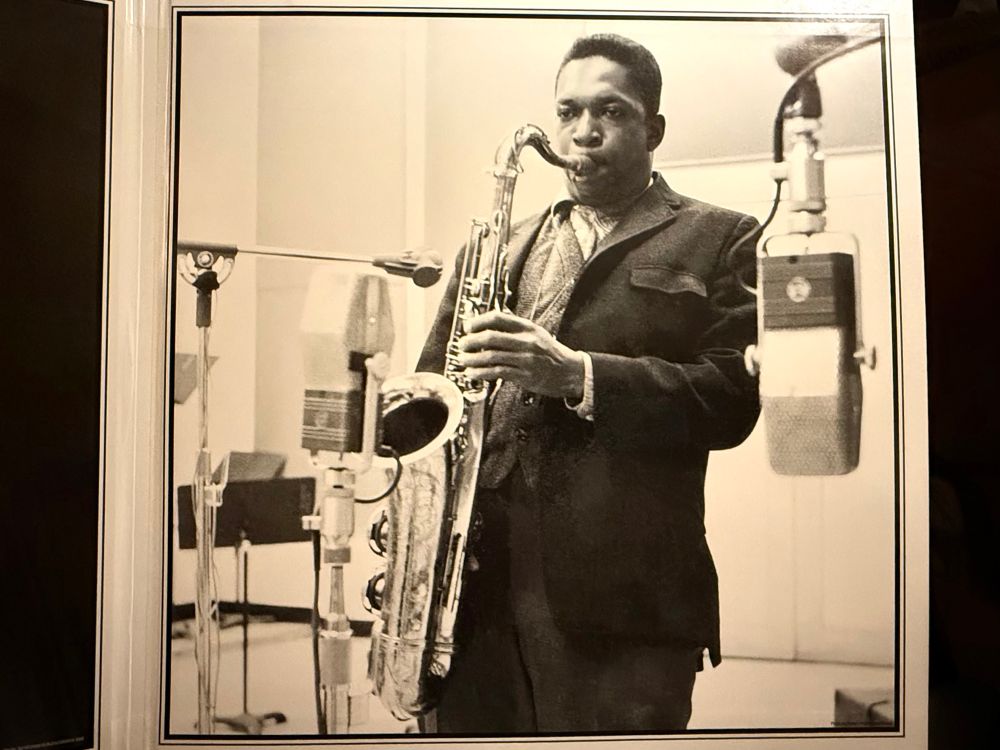 Photo: Robert A. Sengstacke

This black-and-white photo features the legendary jazz saxophonist John Coltrane in a recording studio. He is seen standing, intently playing his saxophone, with his eyes focused ahead, exuding deep concentration. Coltrane is dressed in a dark suit with a light-colored shirt underneath. Surrounding him are large vintage studio microphones positioned to capture his performance. The background is simple, highlighting the professional studio environment, with music stands and additional equipment faintly visible. The overall image captures the essence of a focused artist deeply engaged in his craft.