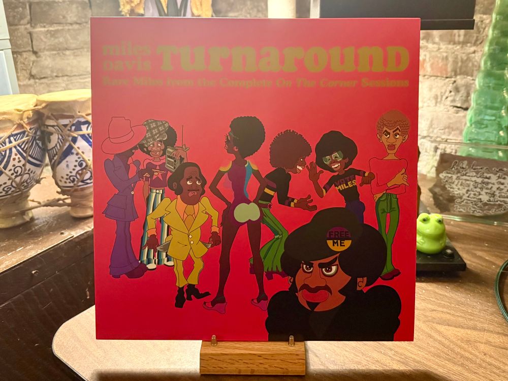 The photo shows an album cover titled "Turnaround: Rare Miles from the Complete On The Corner Sessions" by Miles Davis. The cover features bold, colorful cartoon-style illustrations of various characters dressed in funky, vibrant 1970s attire, including flared pants, platform shoes, and wide-brimmed hats. One character has "MILES" written on their shirt, and another wears a badge that says "FREE ME." The background is bright red, making the colorful characters stand out. The album is displayed upright on a wooden stand, with a rustic brick wall and decorative items like painted drums and a frog figurine in the background.