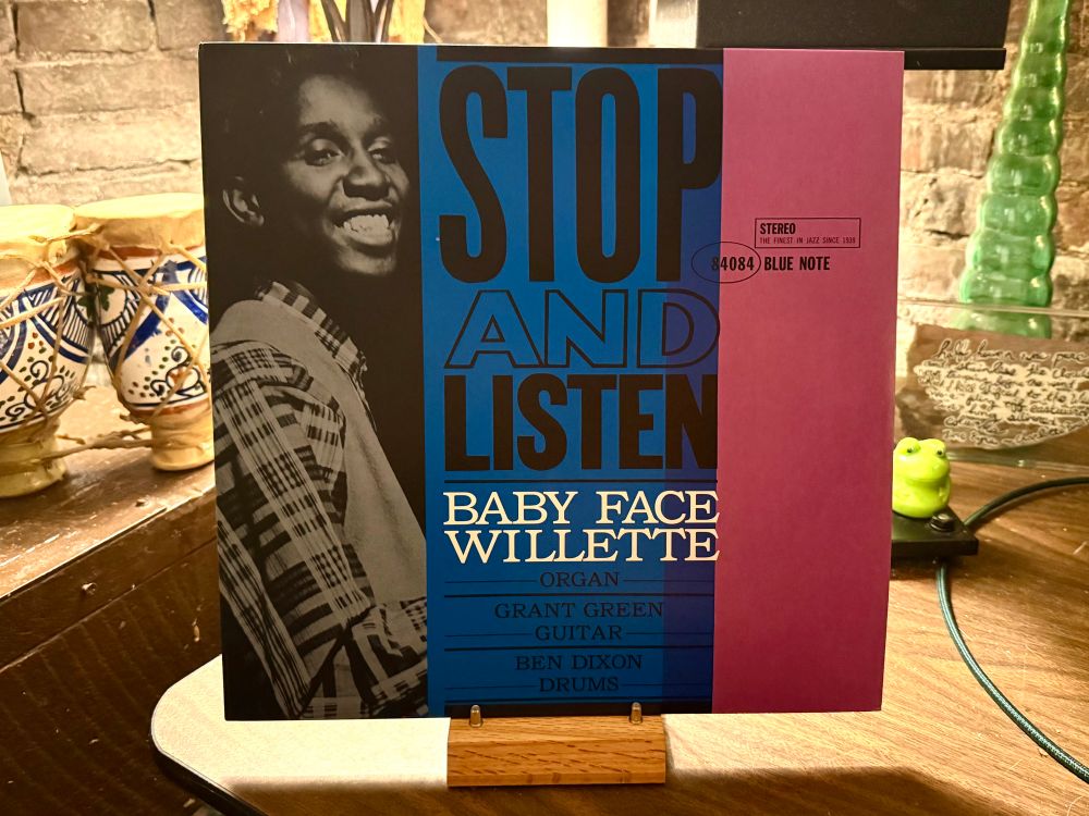 This photo shows a vinyl album cover titled "Stop and Listen" by Baby Face Willette. The cover features a black-and-white photo of Baby Face Willette smiling on the left side. The album title is prominently displayed in large blue and black letters, with the artist's name in white text below. Supporting musicians listed include Grant Green on guitar and Ben Dixon on drums. The album is released under the Blue Note label, indicated by the logo on the right side of the cover. The background includes a cozy setting with decorative ceramic items, a small green frog figurine, a lamp, and handwritten notes on a piece of glass or acrylic. The album is propped up on a wooden stand.