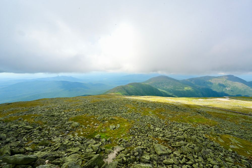 The view from Mount Washington

The photo captures a breathtaking mountain landscape with a rocky foreground covered in patches of green moss and grass. The expansive view stretches to rolling hills and distant peaks, blanketed in shades of blue and green. Soft, diffused light breaks through the thick, low-hanging clouds, casting a gentle glow over parts of the terrain. The vastness of the scene evokes a sense of openness and natural beauty.