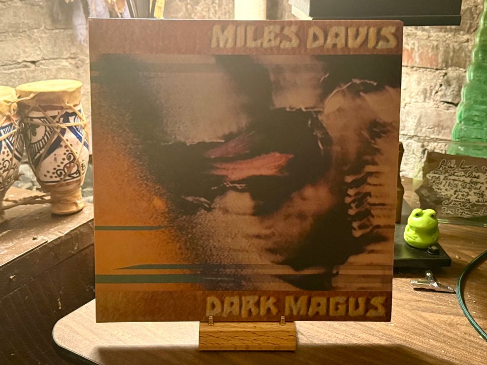 The photo shows a vinyl record cover for "Dark Magus" by Miles Davis. The album cover has an abstract, distorted image of Miles Davis with a mix of dark and warm hues, primarily in shades of brown, orange, and black. The text "MILES DAVIS" is written at the top in bold, yellowish letters, while "DARK MAGUS" appears at the bottom in similar bold, yellow letters. The record is displayed on a wooden stand. In the background, there's a rustic setting with some decorative items including a pair of small, hand-painted drums, a green frog figurine, a green glass bottle, and a handwritten note. The setting appears to be cozy with warm lighting and a brick wall in the background.