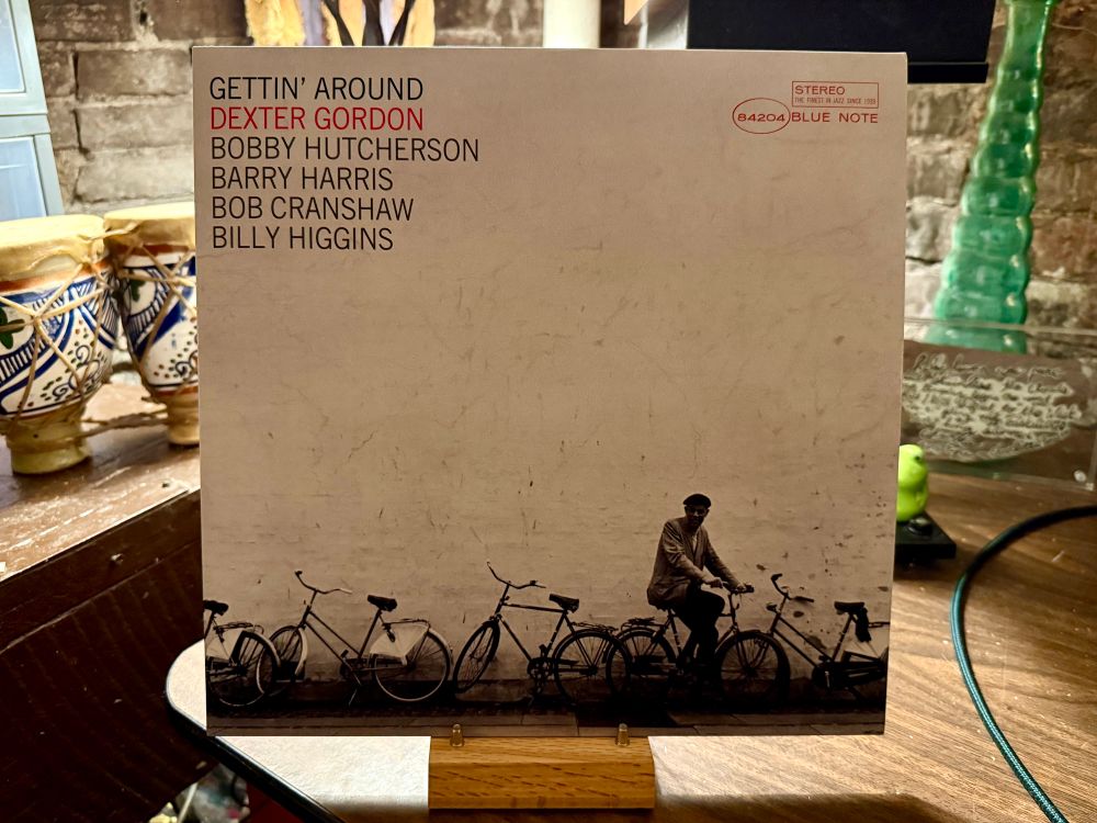 This photo features a vinyl record album cover displayed on a wooden stand. The album is titled "Gettin' Around" by Dexter Gordon, with other musicians listed: Bobby Hutcherson, Barry Harris, Bob Cranshaw, and Billy Higgins. The album is released under the Blue Note label, indicated by the logo and catalog number "84204" in the top right corner.

The cover art is minimalist, showcasing a monochromatic image of a man sitting casually on a bicycle, surrounded by several other bikes, against a plain, textured wall. The setting suggests an urban environment. In the background, there are decorative ceramic items on the left and a green glass bottle on the right, set against a rustic brick wall. The photo is taken in a cozy, eclectic indoor space with warm lighting.