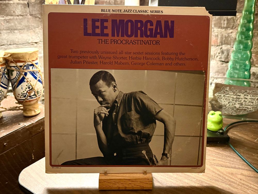 This photo shows the cover of a vinyl record from the Blue Note Jazz Classic Series titled "The Procrastinator" by Lee Morgan. The album cover features a sepia-toned photograph of Lee Morgan sitting thoughtfully with his hand resting on his chin. The text above the photo reads, "Two previously unissued all-star sextet sessions featuring the great trumpeter with Wayne Shorter, Herbie Hancock, Bobby Hutcherson, Julian Priester, Harold Mabern, George Coleman and others." The album cover is placed on a wooden stand, with some decorative items in the background, including patterned pottery, a green glass sculpture, and a small frog figurine.