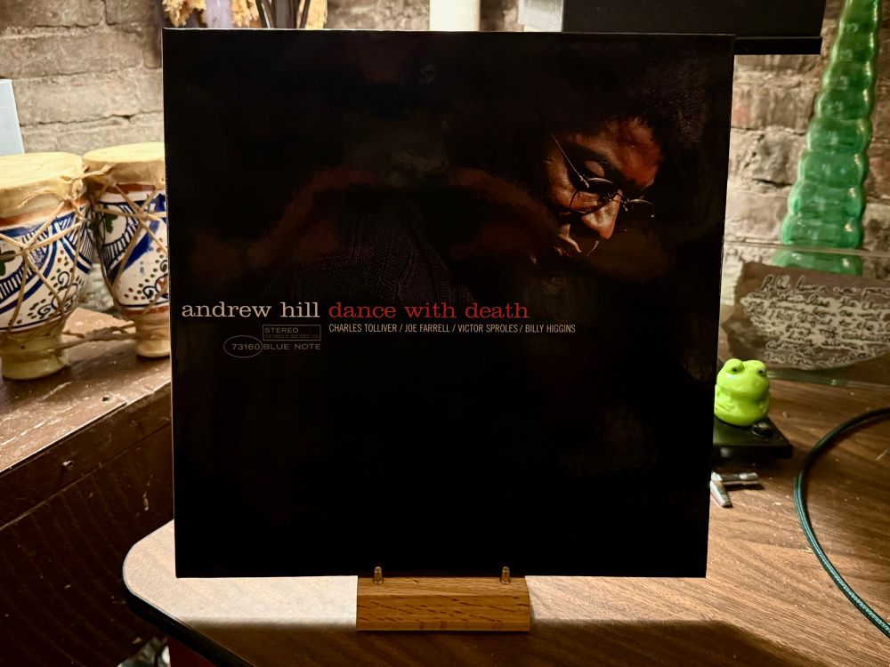 This photo features the album cover of "Dance with Death" by jazz pianist Andrew Hill. The cover has a dark, moody aesthetic with a shadowy image of Andrew Hill, wearing glasses, looking downward. The album title is written in lowercase letters: "andrew hill" in white and "dance with death" in red. Below the title, the names of the contributing musicians—Charles Tolliver, Joe Farrell, Victor Sproles, and Billy Higgins—are listed in small white text. The Blue Note Records logo and catalog number (73160) are also visible. The album is displayed on a wooden stand, set on a table. In the background, there are decorative items, including a pair of hand-painted drums, a small green frog figurine, and a green glass Christmas bottle. The setting appears to be a cozy, eclectic room with a rustic, artistic vibe.