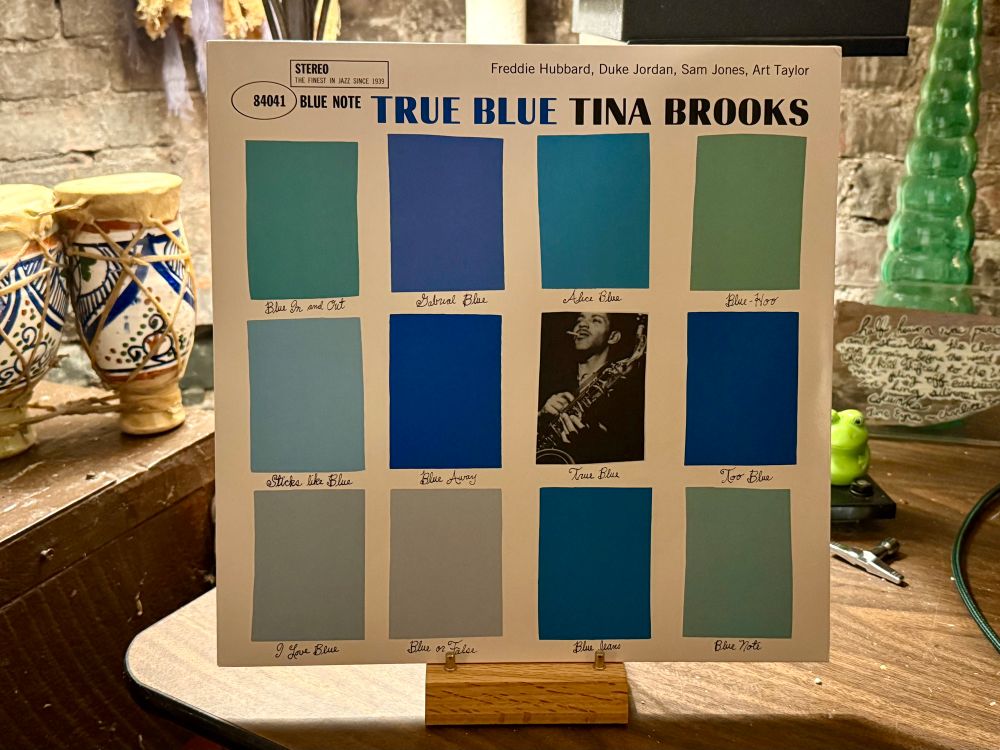 This photo shows the album cover of "True Blue" by Tina Brooks, a Blue Note Records release featuring Freddie Hubbard, Duke Jordan, Sam Jones, and Art Taylor. The cover design has a distinctive grid layout with blue-toned rectangles of varying shades, surrounding a black-and-white photograph of Tina Brooks playing the saxophone. The album title, "TRUE BLUE TINA BROOKS," is prominently displayed across the top in bold blue and black text. The background appears to be a rustic setting with wooden surfaces, decorative pottery with blue patterns, and a brick wall. The album is propped up on a wooden stand.