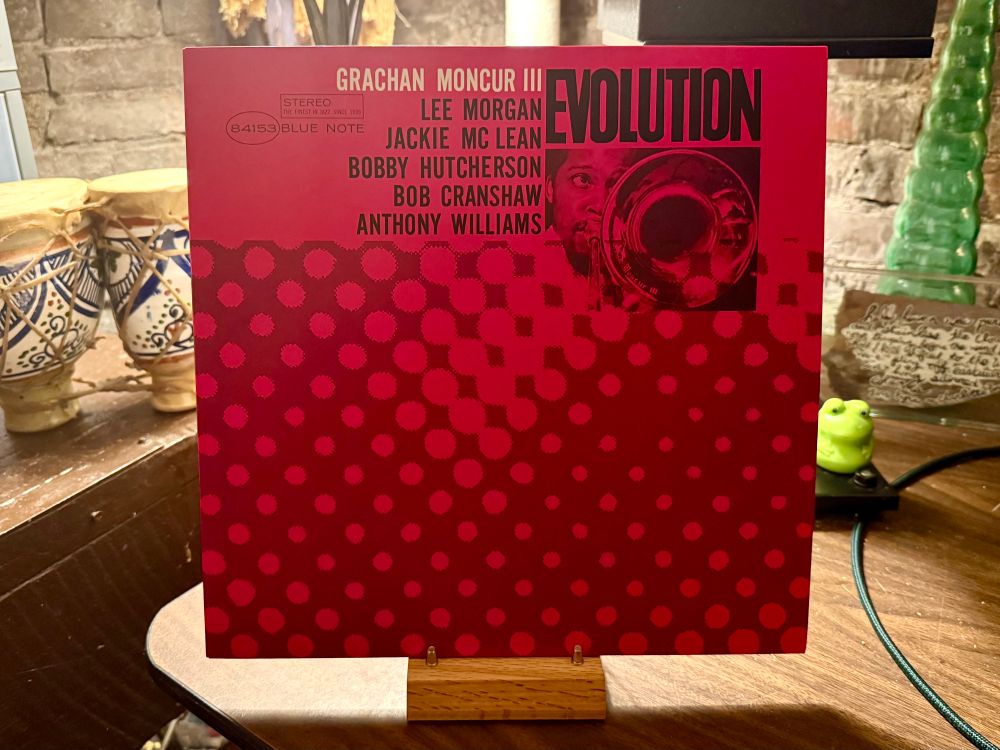 This photo shows the cover of the jazz album "Evolution" by Grachan Moncur III. The album features a striking design with a red color scheme and a pattern of dark red dots fading towards the bottom. In the upper right section, there's a black-and-white photo of a trombone player, likely Grachan Moncur III himself, with the trombone bell prominently visible. The album title "EVOLUTION" is in bold, black uppercase letters. Below Moncur's name, the featured musicians are listed: Lee Morgan, Jackie McLean, Bobby Hutcherson, Bob Cranshaw, and Anthony Williams. The Blue Note Records logo and catalog number "84153" are also visible, indicating its release under the iconic jazz label. The album is displayed upright on a wooden stand, with some decorative items in the background, including ceramic vessels and a small green frog figurine.