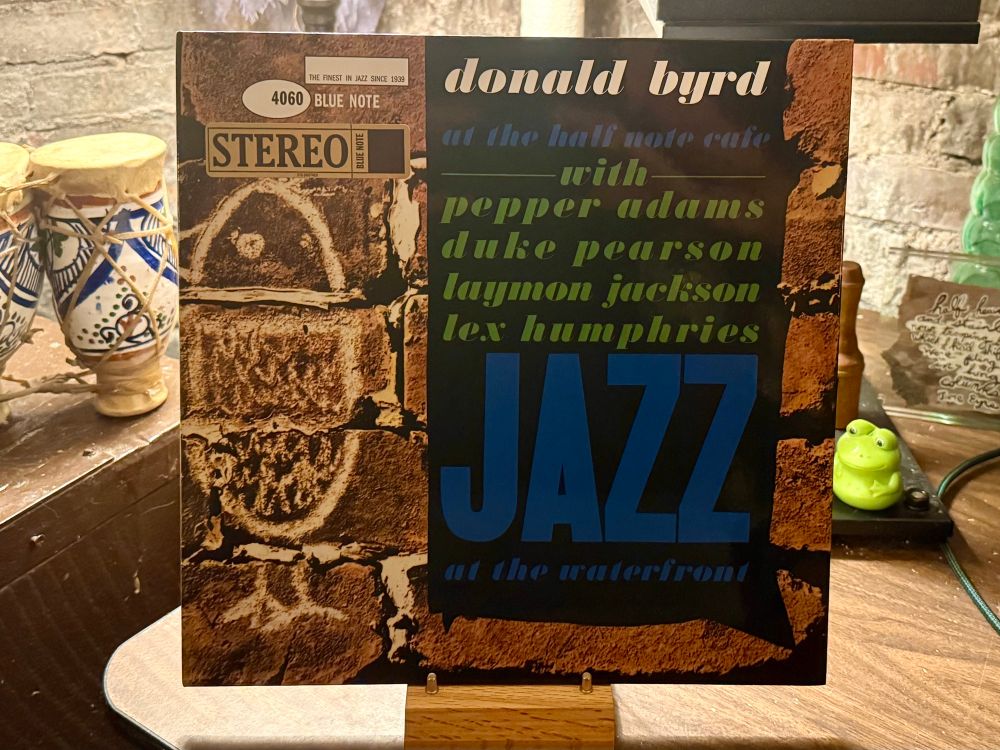 The photo shows the album cover of Donald Byrd's "At the Half Note Cafe" prominently displayed on a wooden stand. The cover features a brick wall background with bold typography in blue and green stating "JAZZ at the waterfront," along with the names of fellow musicians: Pepper Adams, Duke Pearson, Laymon Jackson, and Lex Humphries. The Blue Note Records label is visible at the top left, indicating catalog number 4060 and a "STEREO" label. To the left of the album, there are two decorative ceramic items with blue patterns, possibly vases or cups. On the right, there is a small, green frog figurine, a black gadget with wires, and a note board with cursive writing. The setting appears to be a cozy, rustic space with a brick wall in the background.