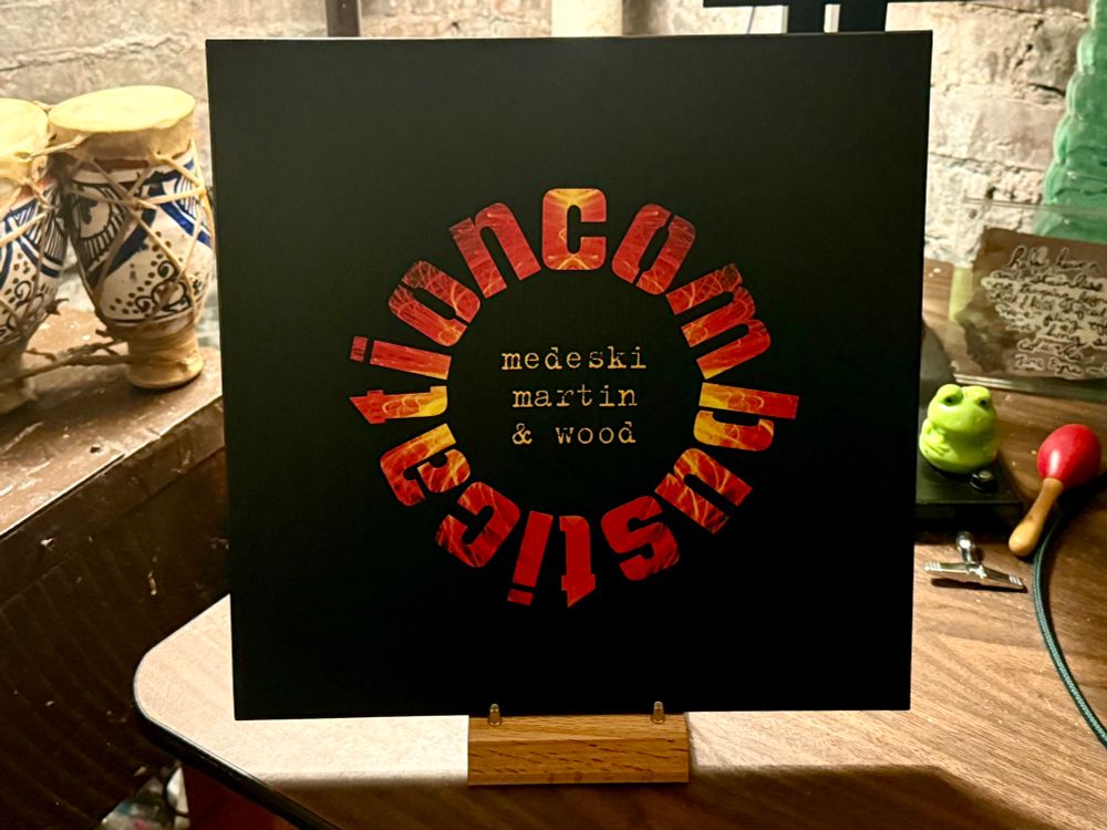 The photo shows a vinyl record cover displayed on a stand. The cover is predominantly black with the word "combustication" in large, bold, gradient lettering that transitions from yellow to red. Below this, the names "medeski martin & wood" are printed in smaller white text. The background includes a wooden surface with eclectic items such as patterned ceramic vases and a frog figurine, adding a cozy, artistic vibe to the scene.