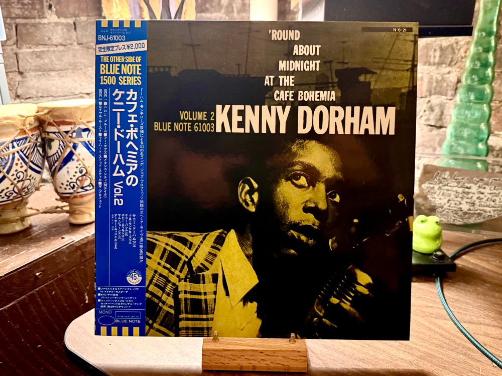 This photo showcases a vinyl record album titled "'Round About Midnight at the Cafe Bohemia, Volume 2" by Kenny Dorham, released under Blue Note Records (catalog number 61003). The album cover features a monochromatic image of Kenny Dorham with a trumpet, predominantly in shades of yellow and black. The left side of the album has an OBI strip with Japanese text, indicating it is a Japanese pressing. The OBI strip mentions "The Other Side of Blue Note 1500 Series" and other details in Japanese, including the price ¥2,000. The record is propped up on a wooden stand, with decorative pottery pieces and a green glass figurine in the background, set on a wooden surface.