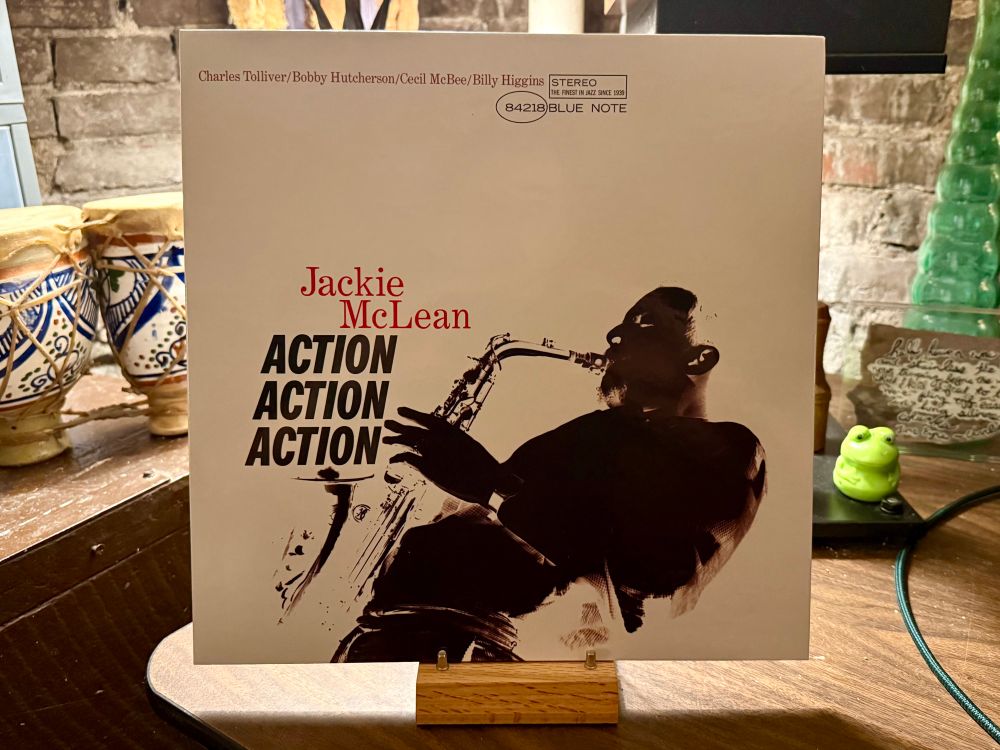 The photo shows the cover of the album "Action Action Action" by Jackie McLean, a renowned jazz saxophonist. The album cover features a striking black-and-white silhouette of McLean playing the saxophone, with bold red and black text highlighting his name and the album title. The cover also lists the names of other musicians featured: Charles Tolliver, Bobby Hutcherson, Cecil McBee, and Billy Higgins. The Blue Note Records logo is visible at the top right. The album is displayed on a wooden stand, with a rustic background that includes some ceramic items and a green frog figurine.