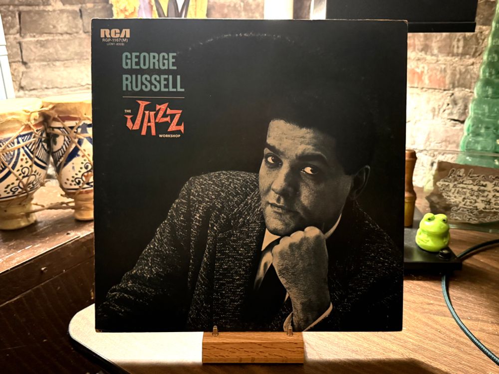This photo features the cover of a vinyl record displayed on a wooden stand. The album is titled "The Jazz Workshop" by George Russell and released under the RCA label (catalog number RGP-1167(M)). The cover has a monochromatic design with a black background and a grayscale portrait of George Russell, who is resting his chin on his hand and looking directly at the camera. The text "GEORGE RUSSELL" is in teal capital letters, while "THE JAZZ WORKSHOP" is in an orange, stylized font that resembles handwritten letters. In the background, there are various decorative items, including patterned ceramic vases and a small green frog figurine placed on a dark surface, along with a brick wall partially visible.