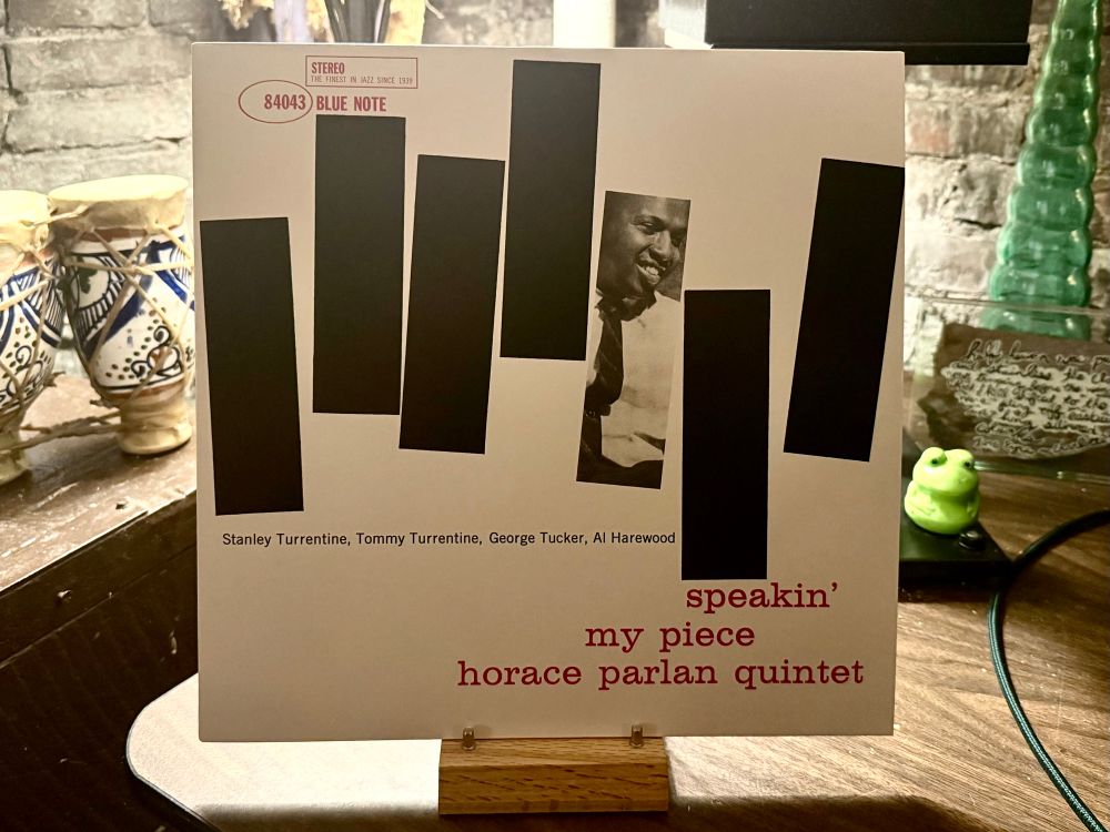 This photo features the cover of a vinyl record album titled "Speakin' My Piece" by the Horace Parlan Quintet. The album is a release from the Blue Note label, catalog number 84043, as indicated in the top left corner. The cover design consists of bold, black vertical rectangles arranged in a staggered fashion, resembling piano keys, with a black-and-white photo of Horace Parlan integrated into one of the rectangles. The names of the quintet members—Stanley Turrentine, Tommy Turrentine, George Tucker, and Al Harewood—are listed below the rectangles. The background is a cozy setting with decorative pottery, a frog figurine, and other eclectic items placed on a wooden surface.