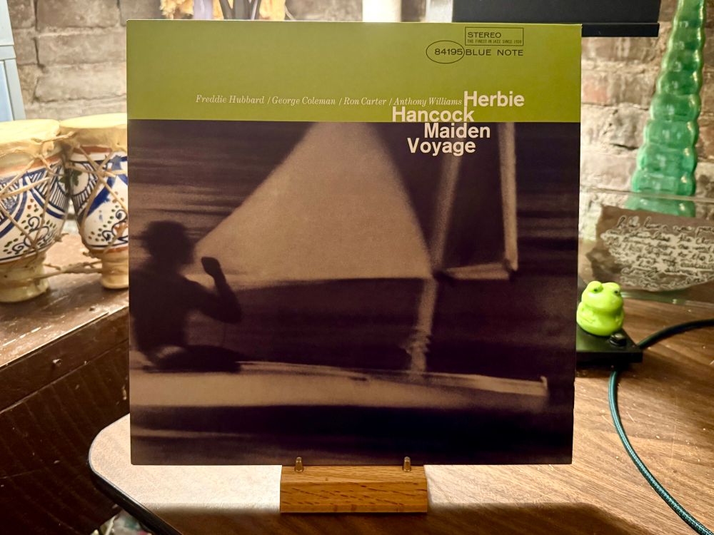 This photo features the cover of the jazz album "Maiden Voyage" by Herbie Hancock, released under the Blue Note label. The album cover is displayed upright on a wooden stand. The cover design has a green band at the top with the names of the musicians—Freddie Hubbard, George Coleman, Ron Carter, and Anthony Williams—along with Herbie Hancock’s name prominently featured. Below, there's a blurred, artistic image of a person sailing, evoking a sense of motion and water.

In the background, the setting appears to be a cozy indoor space with various decorative items. On the left, there are two traditional ceramic drums with intricate blue designs. To the right, there is a small green frog figurine and a handwritten note or card. A green glass decorative object is seen further back, and the table surface has a wooden texture with some cables visible.