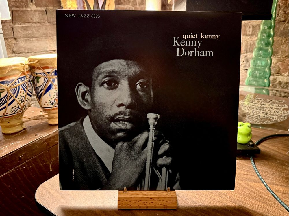 The photo shows a vinyl record cover for the jazz album Quiet Kenny by Kenny Dorham. The cover is mostly black with a black-and-white portrait of Kenny Dorham holding a trumpet. The text reads "NEW JAZZ 8225" at the top left and “quiet kenny Kenny Dorham” in white on the upper right. The record is propped on a wooden stand on a wooden surface. In the background, there are ceramic drums with colorful patterns, a green frog figurine, a translucent green lamp, and what appears to be a handwritten note on a clear surface. The setting has a cozy, rustic feel with a brick wall visible in the background.