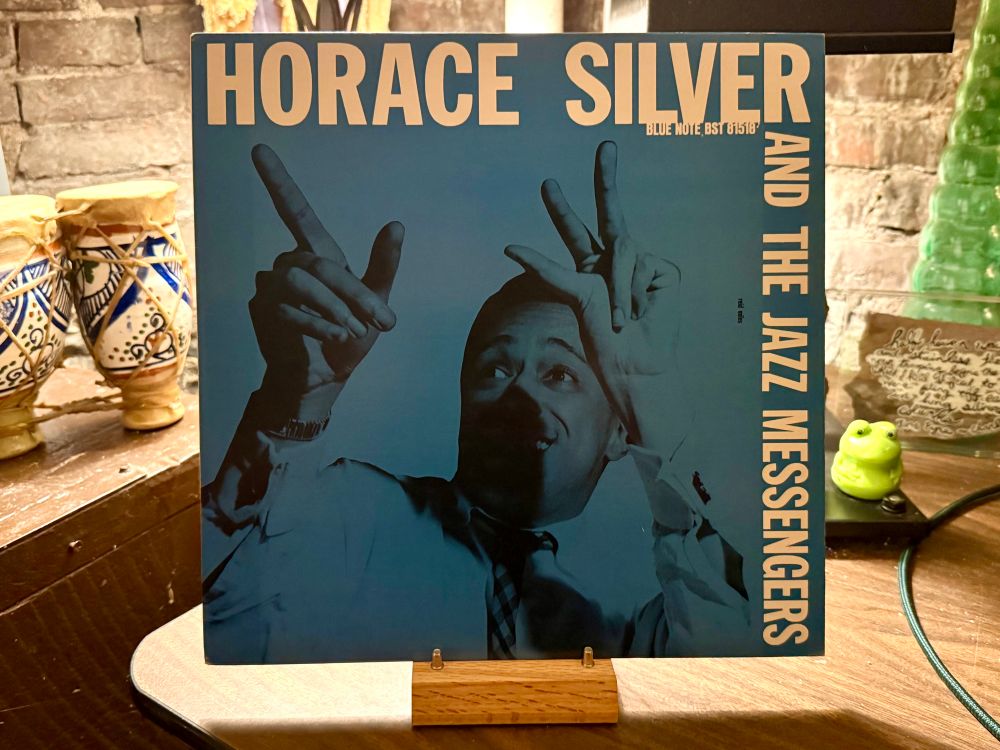 The photo features a vinyl record titled "Horace Silver and The Jazz Messengers" displayed upright on a wooden stand. The album cover has a blue-tinted image of Horace Silver, who is gesturing with both hands, pointing upward with an animated expression. The text on the cover is bold and capitalized, with "HORACE SILVER" prominently displayed at the top in large beige letters. The background includes some decorative ceramic items on the left, a small green frog figurine on the right, and a note with cursive handwriting partially visible. The setting appears to be a cozy indoor space with a rustic textured wall in the background.