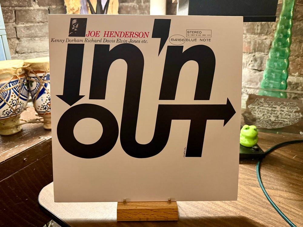 The photo shows the album cover of "In 'n Out" by jazz saxophonist Joe Henderson. The cover features bold, black typography with the words "in" and "out" stylized using large arrows pointing downward and to the right, respectively. Above the title, Joe Henderson's name is printed in red, along with the names of other contributing artists: Kenny Dorham, Richard Davis, and Elvin Jones. The album is labeled as a Blue Note release, with the catalog number 84166. The background setting includes decorative items like blue-and-white ceramic drums, a green frog figurine, and a handwritten note on glass, all placed on a wooden surface against a brick wall backdrop.