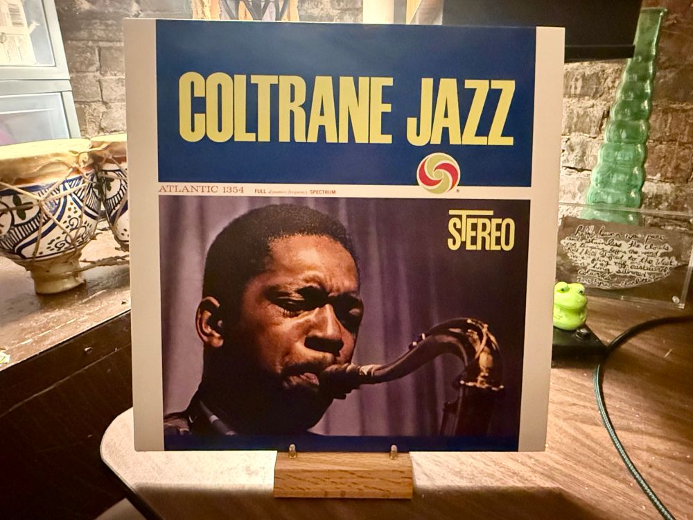 This photo features the album cover of "Coltrane Jazz" by John Coltrane, prominently displayed on a wooden stand. The album cover has a bold blue background at the top with large yellow text reading "COLTRANE JAZZ" and a colorful swirl logo beneath it. Below the title section is a photograph of John Coltrane playing the saxophone, captured in a focused and intense expression. The photo is bordered by text indicating "ATLANTIC 1354" and "STEREO" on the right-hand side. The background includes eclectic decor such as a ceramic bowl with blue patterns, a green glass ornament, and a partially visible handwritten plaque with cursive text. The setting appears cozy, possibly in a cafe or a personal space with warm lighting and a rustic aesthetic.