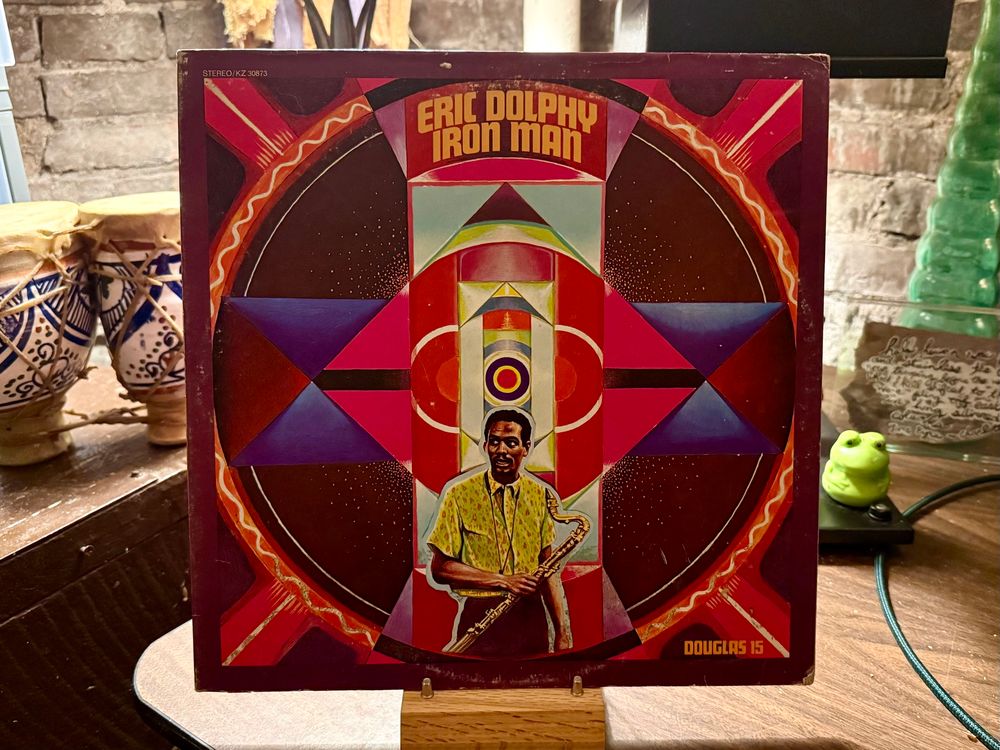 This photo showcases the album cover of "Iron Man" by Eric Dolphy. The cover features a vibrant and abstract geometric design with bold, contrasting colors including red, purple, and black. At the center sits an illustrated portrait of Eric Dolphy holding a saxophone, framed within an intricate pattern of shapes and concentric circles. The album title "ERIC DOLPHY IRON MAN" is displayed prominently at the top in bold, yellow letters. The album appears to be placed on a stand with various decorative items in the background, including ceramic drums on the left and a green frog figure on the right.