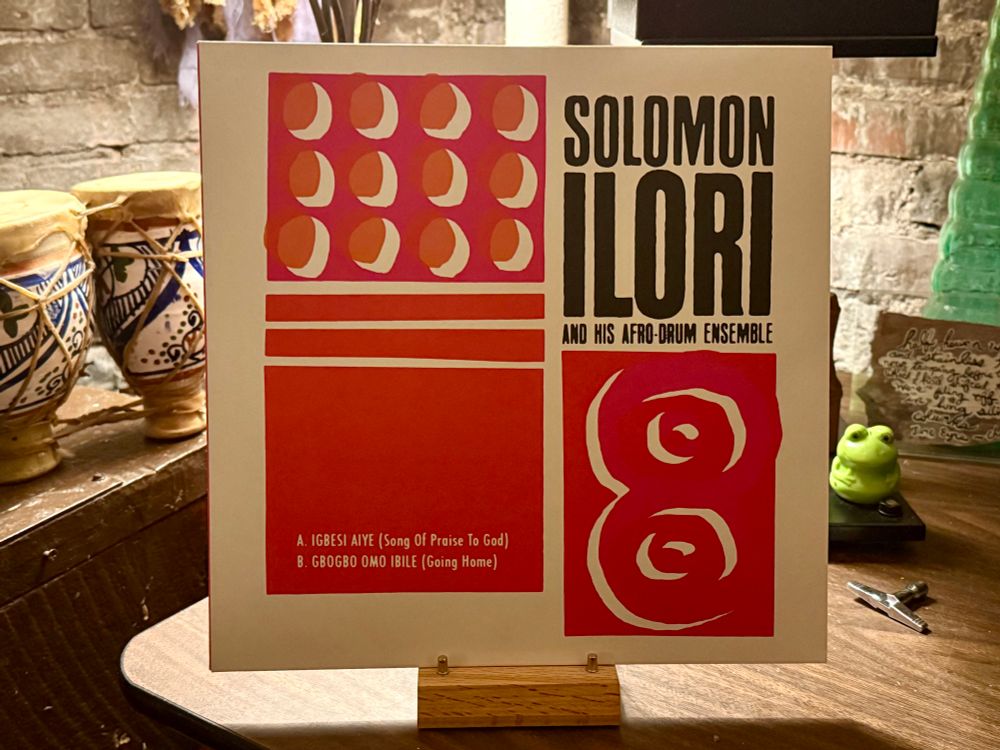 The photo shows an album cover for "Solomon Ilori and His Afro-Drum Ensemble." The design features a bold red and white color scheme with geometric shapes and patterns, including circles and rectangles. The track titles are listed as "A. Igbesi Aiye (Song of Praise to God)" and "B. Gbogbo Omo Ibile (Going Home)." The album is displayed on a wooden stand, with traditional drums and a green frog ornament in the background, set against a brick wall.