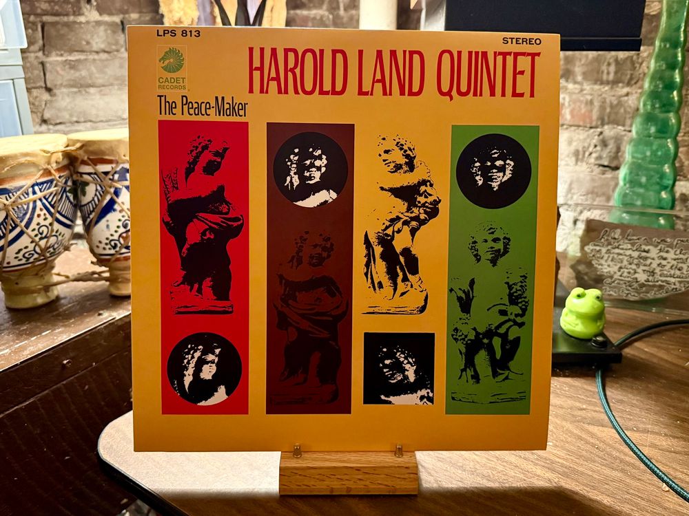 This photo features a vinyl record cover of "The Peace-Maker" by the Harold Land Quintet, released by Cadet Records (catalog number LPS 813). The cover is dominated by yellow with bold red text for the artist's name at the top. The design includes three vertical panels in red, brown, and green, each containing artistic black-and-white images resembling classical sculptures or busts. The record is displayed upright on a small wooden stand. In the background, there's a rustic setting with ceramic pots featuring blue patterns on the left, a green glass decorative piece on the right, and a small green frog figurine next to it. The backdrop includes a brick wall, adding a vintage, cozy ambiance to the scene.