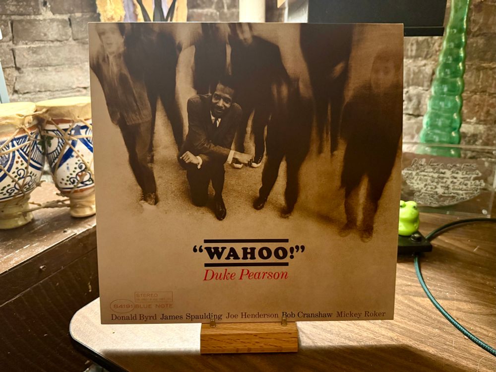 The photo shows the cover of a vinyl record album titled "Wahoo!" by Duke Pearson. The cover features a sepia-toned image of Duke Pearson kneeling and smiling at the camera, surrounded by blurred figures standing around him, creating a dynamic effect. The album features other musicians: Donald Byrd, James Spaulding, Joe Henderson, Bob Cranshaw, and Mickey Roker, listed at the bottom. The record is displayed on a wooden stand on a table. In the background, there are decorative ceramic items, a green glass object, and a brick wall, giving the setting a cozy and eclectic feel.