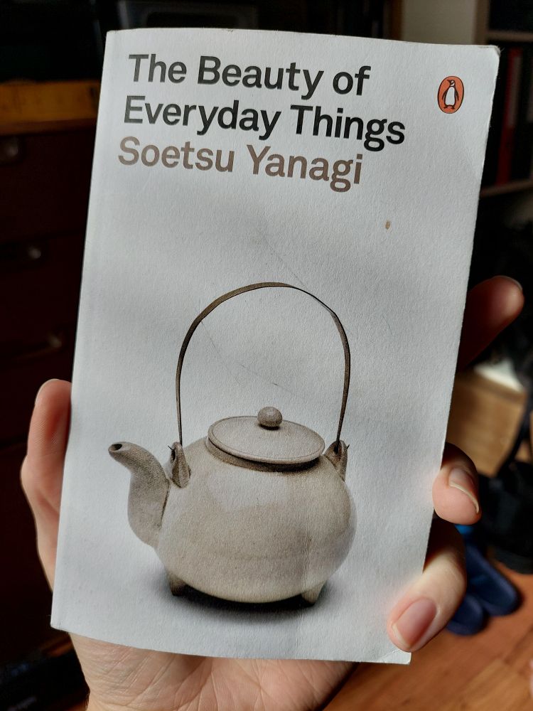 The image shows a hand holding a book titled "The Beauty of Everyday Things" by Soetsu Yanagi. The cover design is minimalist and elegant, featuring a traditional Japanese teapot with a gracefully curved handle and spout—symbolizing the book’s celebration of functional artistry in daily life. In the top right corner, the classic Penguin Books logo is visible, giving it a timeless literary feel.

The background is softly blurred, hinting at a cozy indoor space with furniture and objects that suggest a serene, lived-in atmosphere. The focus on the hand and the book evokes a quiet moment of reflection or curiosity.

It’s a beautiful capture of someone engaging with a work that explores how even the most ordinary things—if crafted with care—can hold deep aesthetic and cultural value.