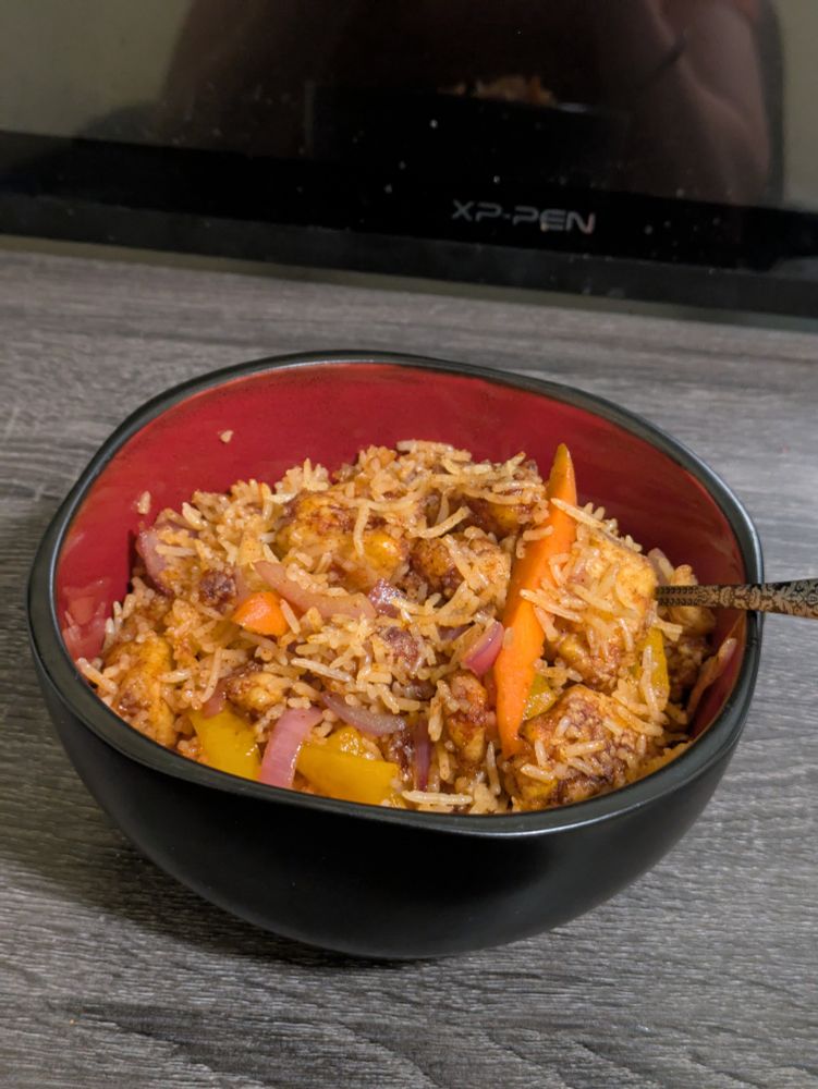 A picture of a black and red bowl sitting on a grey desk. In the bowl is a stir fry, with chunks of pepper, tofu, carrots, and onions visible.