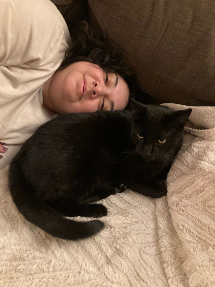 Sarah is laying on a couch and smiling next to Panther, a black cat, who is curled up on a blanket