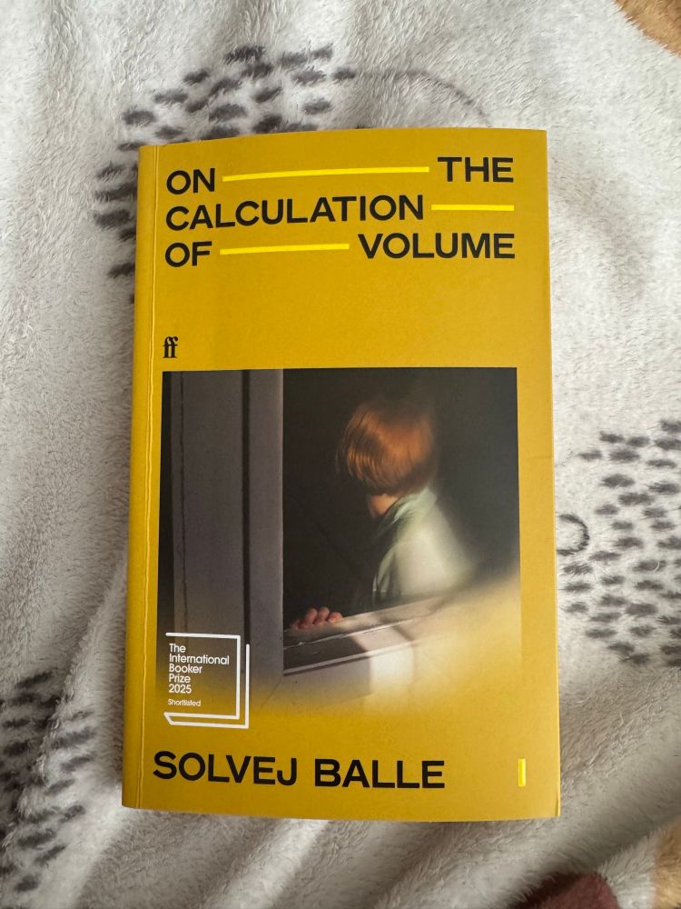 A yellow book cover with black text reading “On the Calculation of Volume” by Solvej Balle. The title words “THE”, “CALCULATION”, and “VOLUME” are underlined. The center of the cover features a photo of a person with short reddish hair, wearing a green top, looking out a window. 