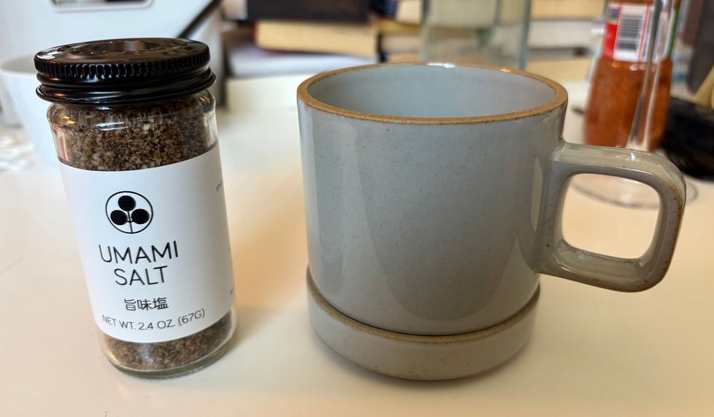 A glossy gray mug and saucer, both made with precise, straight lines. They sit next to a small glass jar of "Umami Salt," which has a brown color from the dried mushroom and black garlic in it.