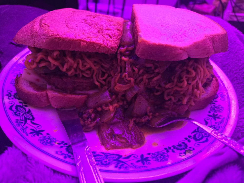 a hamburger on a plate with flowery accents with fork and butter knife stuck under the burgers. plate is sat on the cover im under. screen is purply pink cause of an off screen lamp light. its 4 sheets of white bread one being a heel. bbq sauce, pepper jack cheese, cheap 80 cent patty, pickles, ramen, onion, and thousand island dressing.