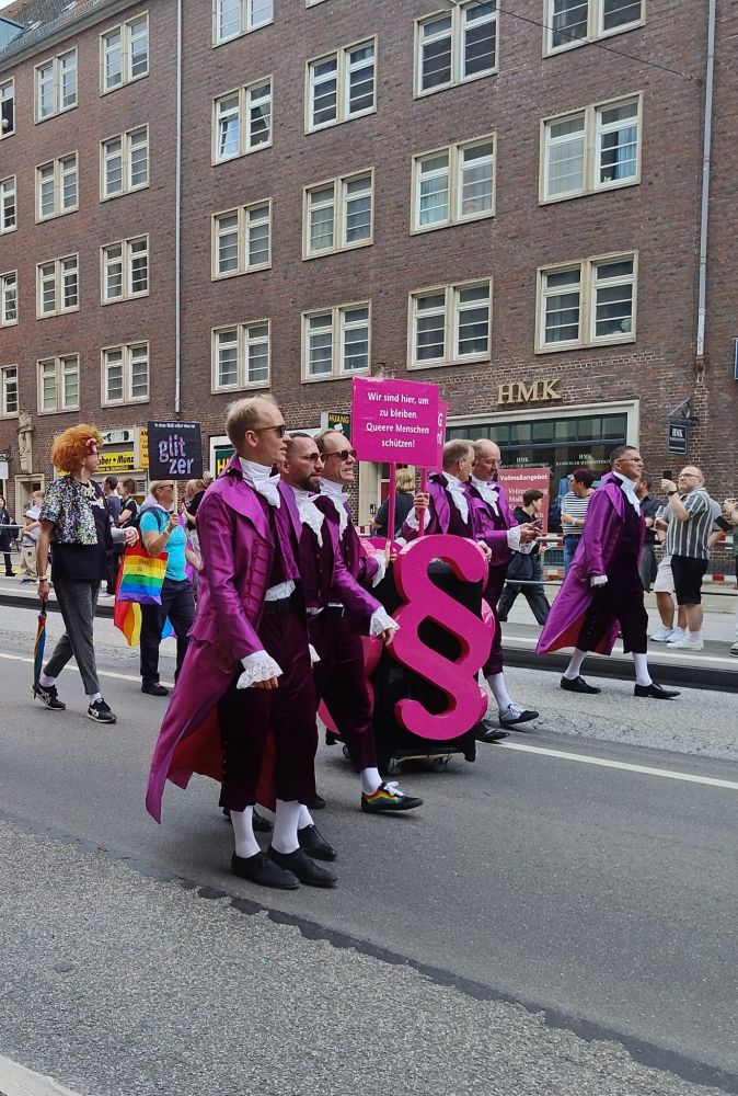 Foto vom CSD Hamburg das pink gekleidete Juristen mit einem xxl pinken Paragraphenzeichen zeigt. 