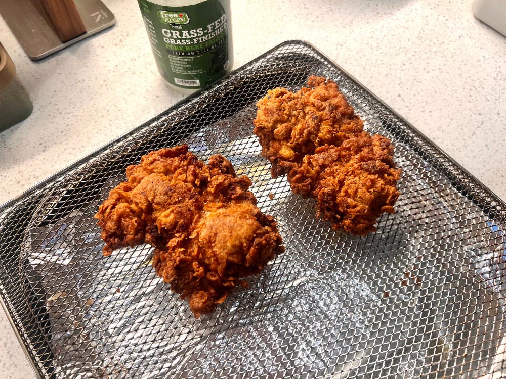 some fresh fried chicken thighs cooling on a rack. there’s a jar of beef tallow in the background 