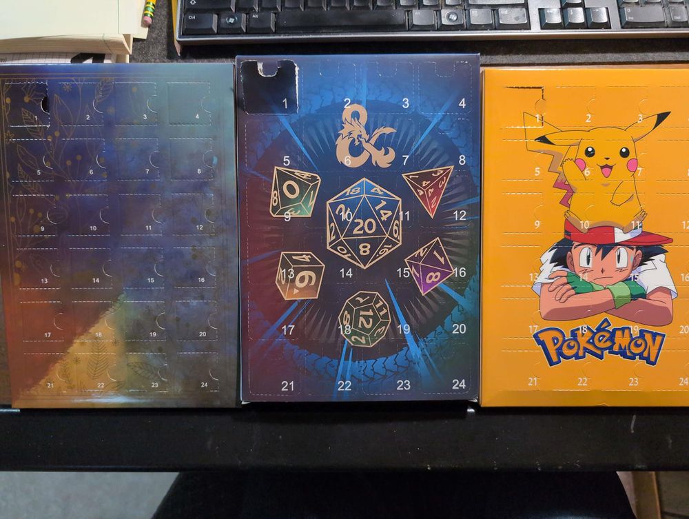 Three advent calendars in a row. The first depicts a gold-colored design with snowflakes, holly, and leaves in gold on a dark colored gradient background. The center has a gold ampersand and polyhedral dice with gold borders. The third depicts Ash Ketchum with an embarrassed expression, slumped with his arms crossed. Pikachu sits on his head, waving at the viewer.