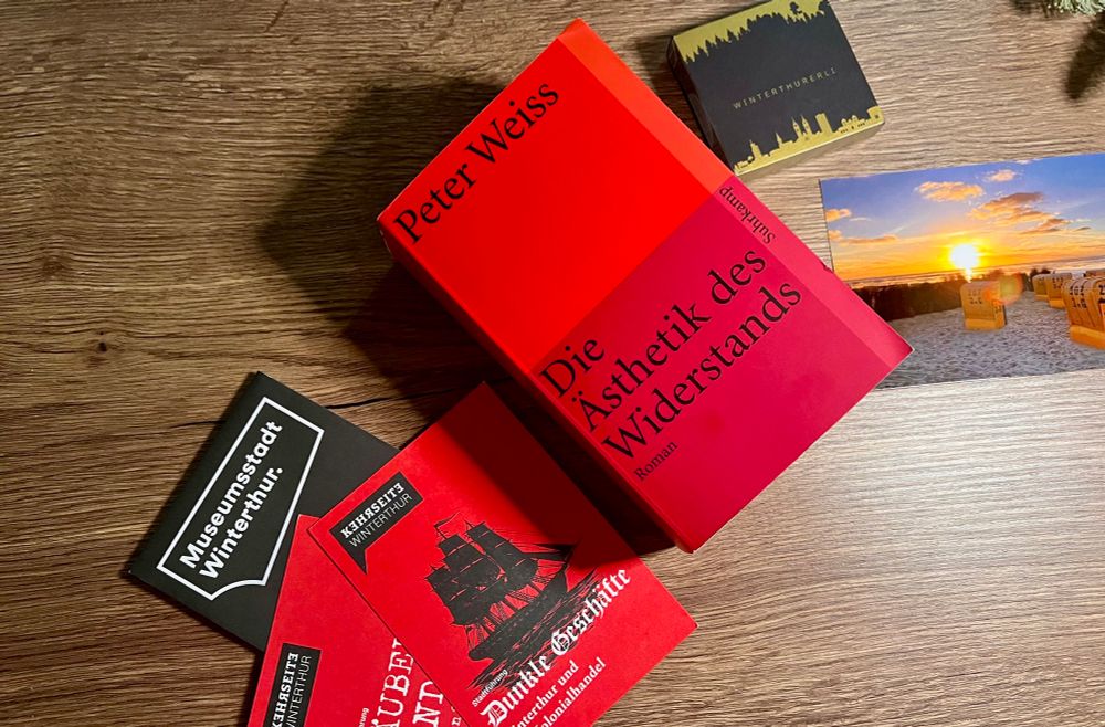 Buch „Die Ästhetik des Widerstands“ von Peter Weiss, links neben dem Buch eine Karte mit Museen in Winterthur, Ansichtskarten, die auf Stadtführungen in Winterthur hinweisen, darunter zur kolonialen Geschichte der Stadt, rechts neben dem Buch eine Ansichtskarte mit einer Strandszene aus Norddeutschland und eine Päckchen Süßigkeiten.