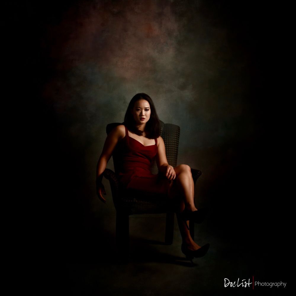 Beautiful Chinese woman of around 30 years of age wearing a dark red dress and sitting in a black and gold chair. Legs crossed.