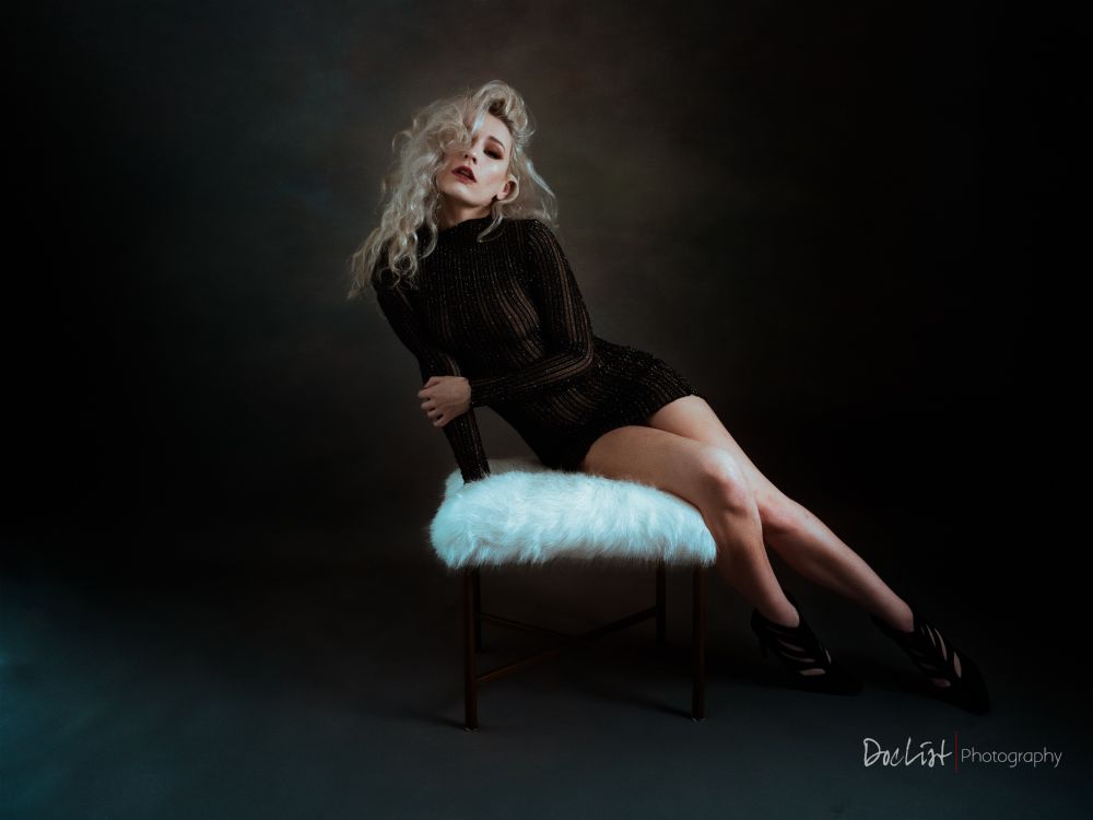 beautiful sultry blonde with wavy hair wearing an almost-see-through black dress and sitting on a furry white ottoman