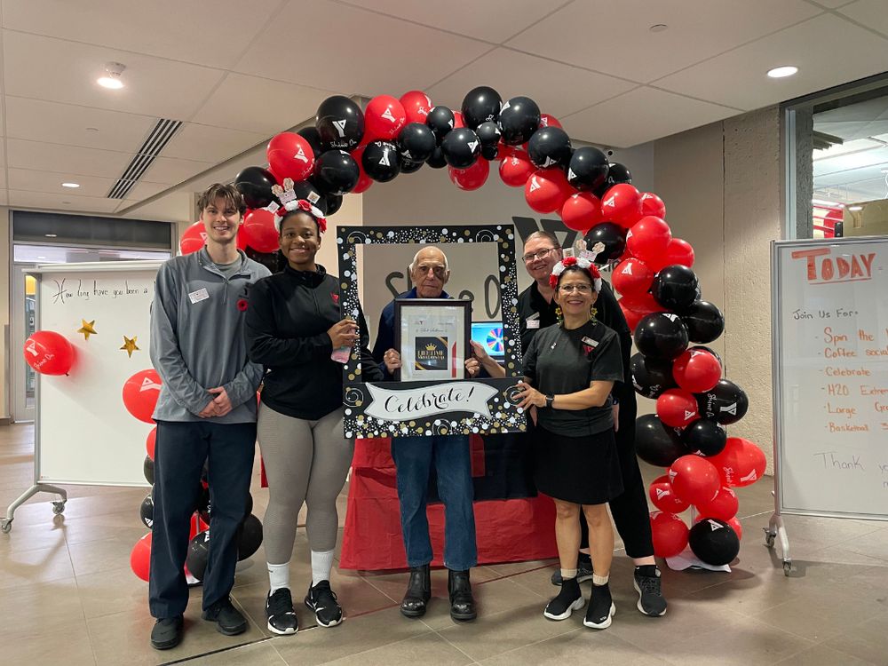 Four Y staff members standing beside a Y member receiving a lifetime award. The staff are holding a frame around him that reads "Celebrate!" along the bottom. They are standing beneath a red and black balloon arch.