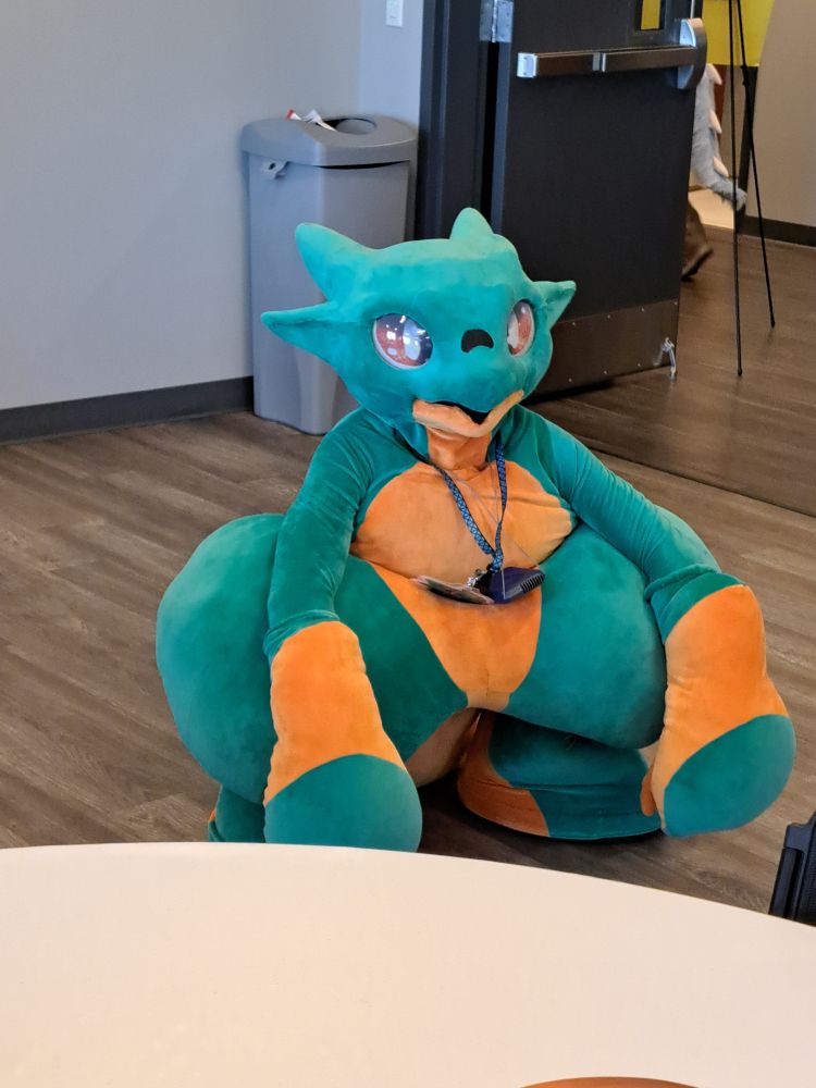 Karudamon, a green dragon fursuit with orange patches on the belly, hands, feet, and tail, sitting on the floor.