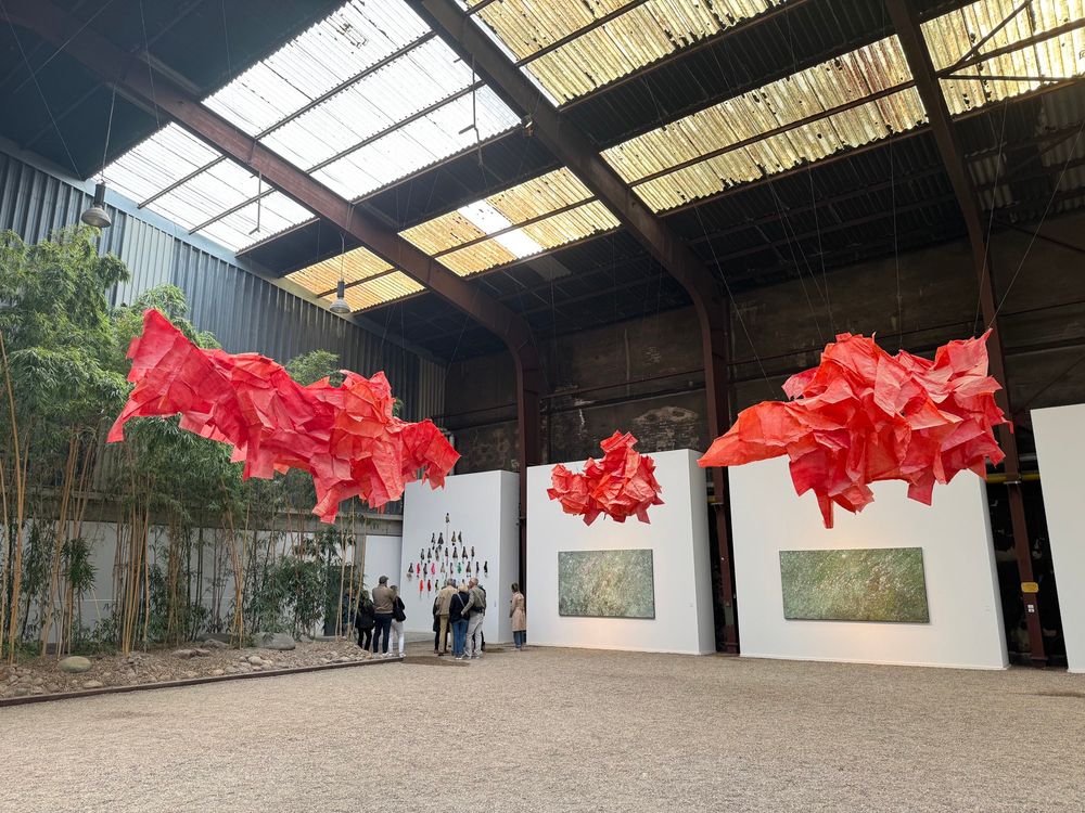 Innenansicht der NordArt-Industriehalle: Von der Decke hängen drei große, wolkenförmige Skulpturen aus rotem, gefaltetem Papier. 