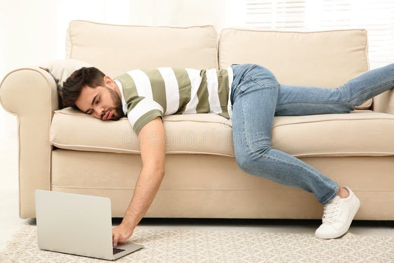 A man lying face-down on a couch typing on a laptop on the floor. His pelvis covers the crack between couch cushions and left leg is poised for a forward thrust.
