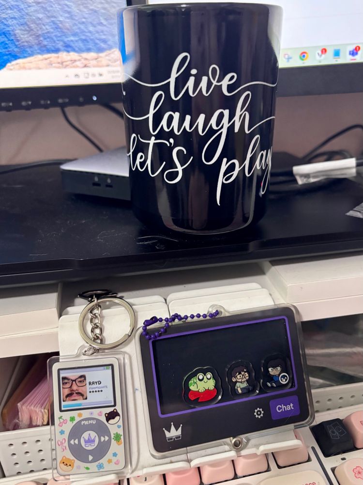 a black mug with live laugh let’s play written in white script and two keychains, one looks like a twitch chat box with emotes in it and the other looks like a classic ipod covered in stickers