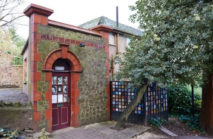 The front exterior of a small, brick building with a dark red half glazed door. There is a holly tree to the right of the building