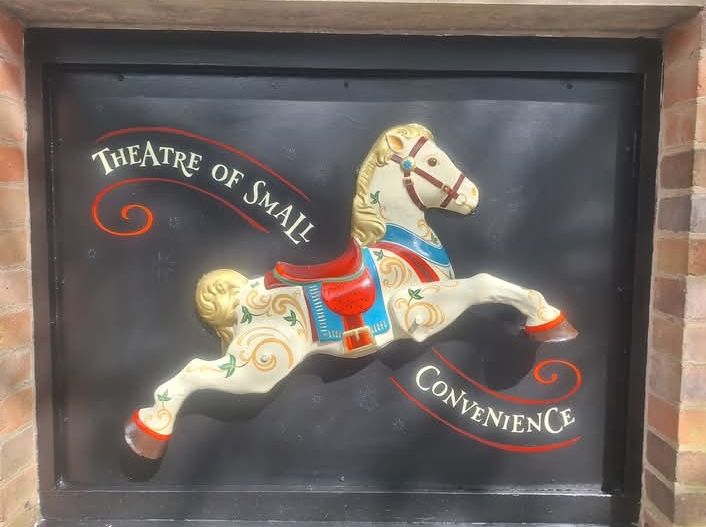 One half of a 1970s metal rocking horse, sawn along the centreline, painted in Victorian fairground style and fixed to a wall on a black background. Decorative text swirls around, reading "Theatre of Small Convenience"