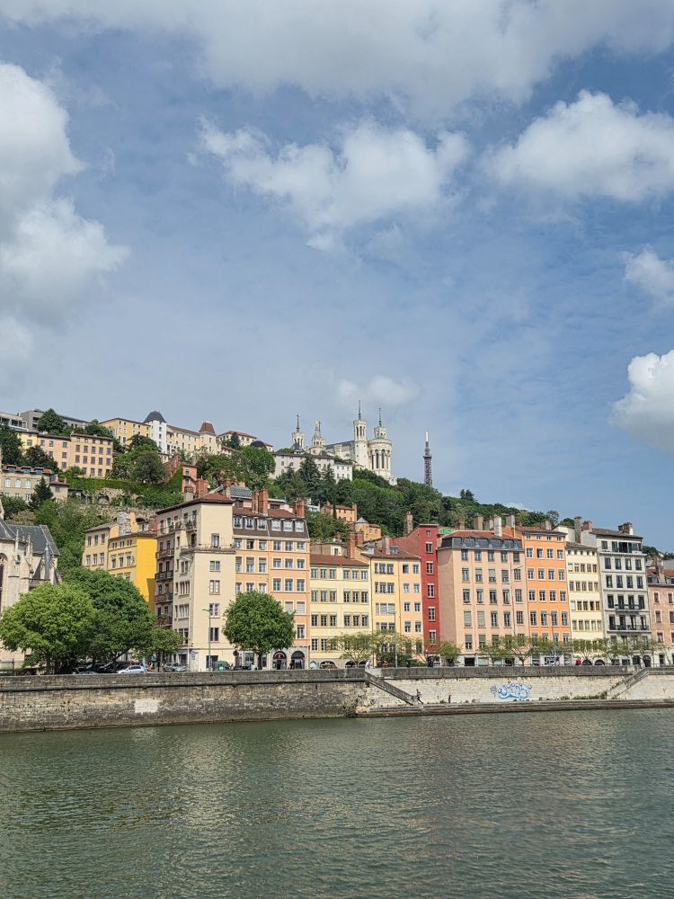 A city scene. Coloured buildings alongside a river. A church at the top of a hill. Blue sky with small clouds