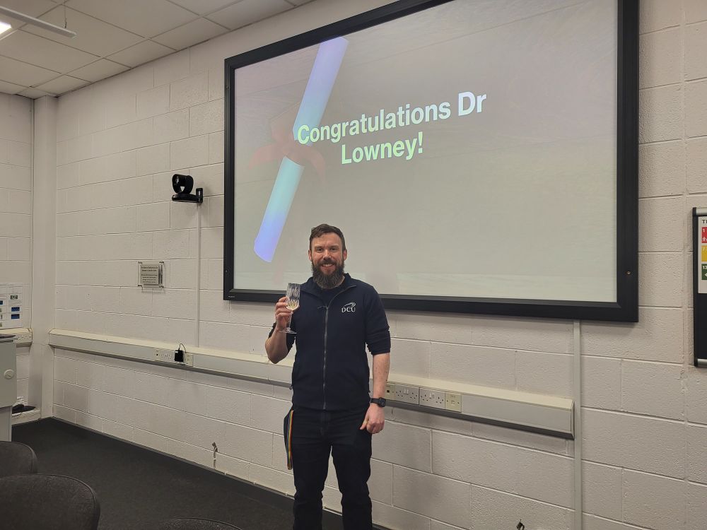 Man standing with glass of champagne in front of a screen with the words congratulations Dr Lowey