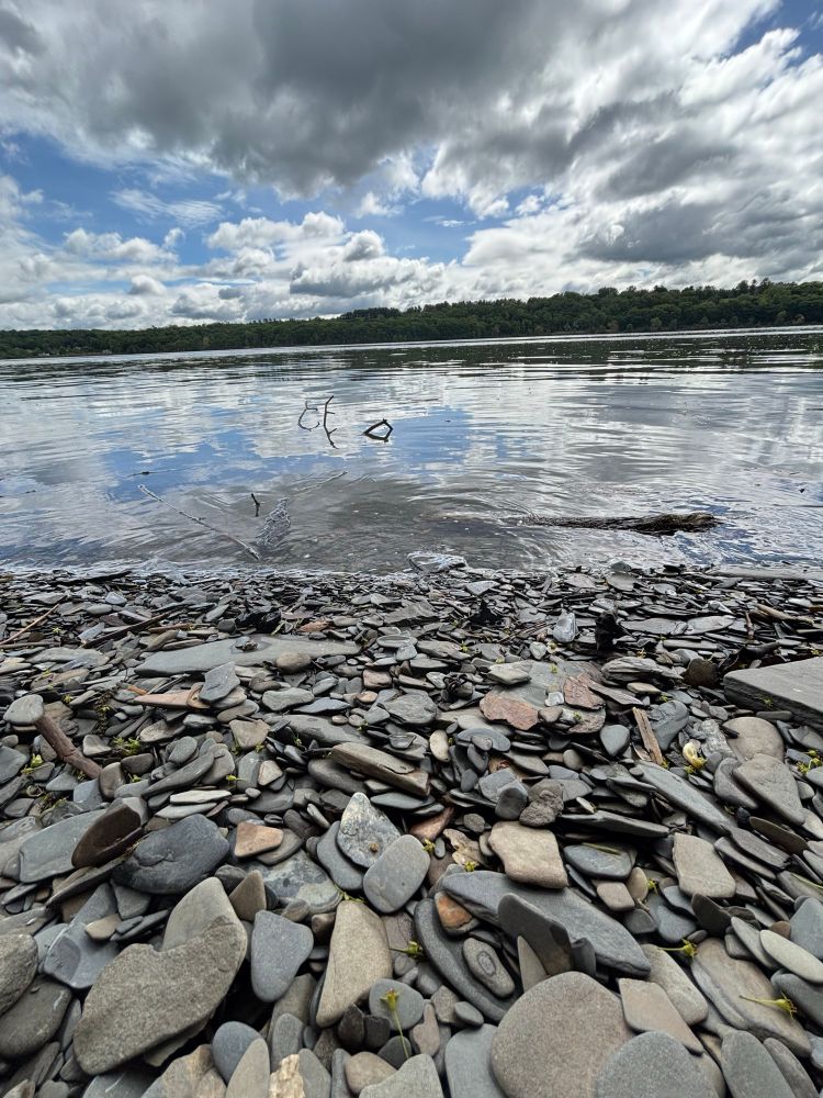 A view of the Hudson River, with clouds above reflected below. Flat stones are in the foreground, tempting one to try skipping them on the water. 