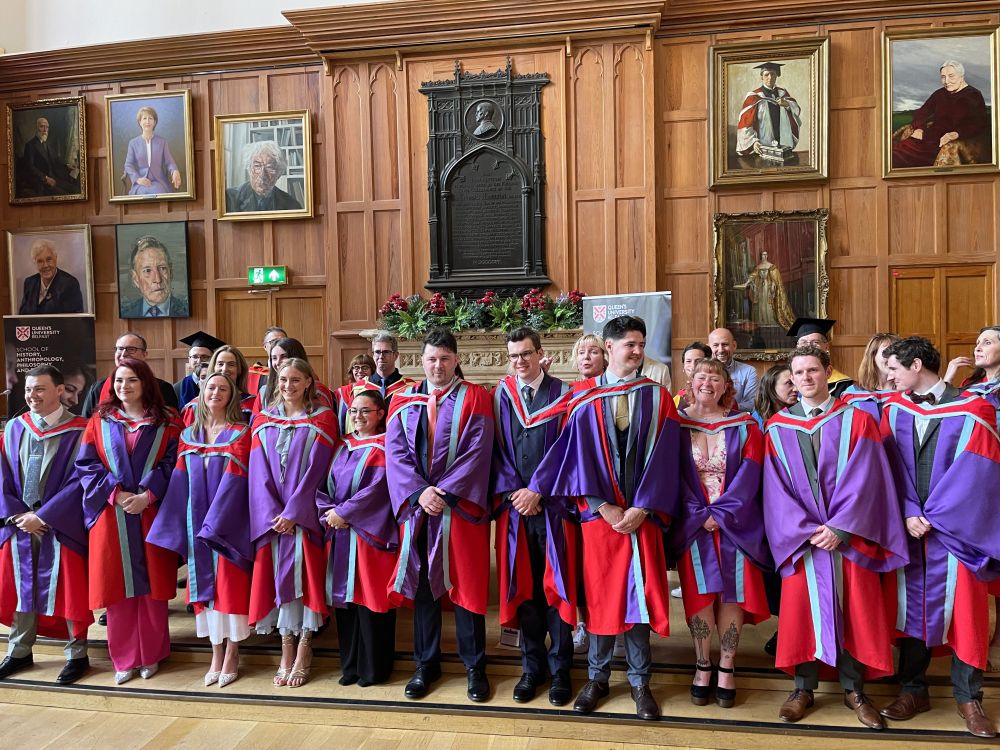 A group of new PhDs in QUB robes standing in the Great Hall, with supervisors behind