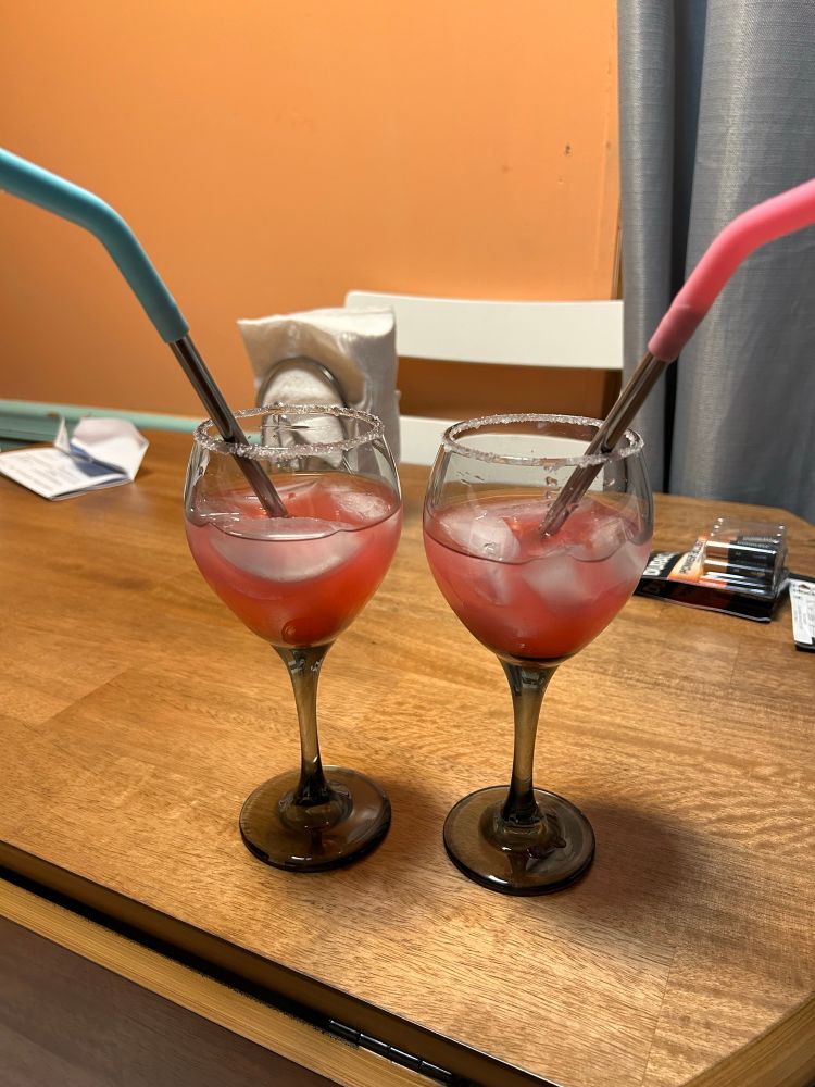 Two glasses sitting on a table filled with margarita wine, they're rimmed with salt/sugar and each have a metal straw inside, topped with a pink/blue silicon piece.