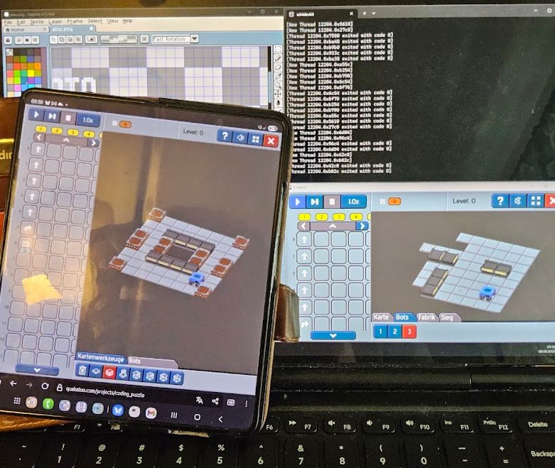 The image shows a development setup for the game 8-bitBot, with multiple devices and screens displaying the game and related tools.

In the foreground, a foldable smartphone is running the game in editor mode. The screen shows the programming panel on the left and an isometric game world on the right, where a robot stands inside a square arena surrounded by conveyor belts and blocks. At the bottom, the “Kartenwerkzeuge” and “Bots” tabs are visible, indicating the map editing mode.

In the background, a laptop screen displays several windows. On the right side, the game is also running in editor mode, showing a different level layout with conveyors and walls. Above it, a black console window from w64devkit shows runtime logs and thread exit codes. On the left side of the laptop screen, a pixel art editor is open, displaying part of a game asset in a zoomed-in grid view.